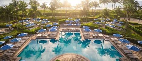 A pool area with lounge chairs and umbrellas, surrounded by palm trees and a view of the ocean.