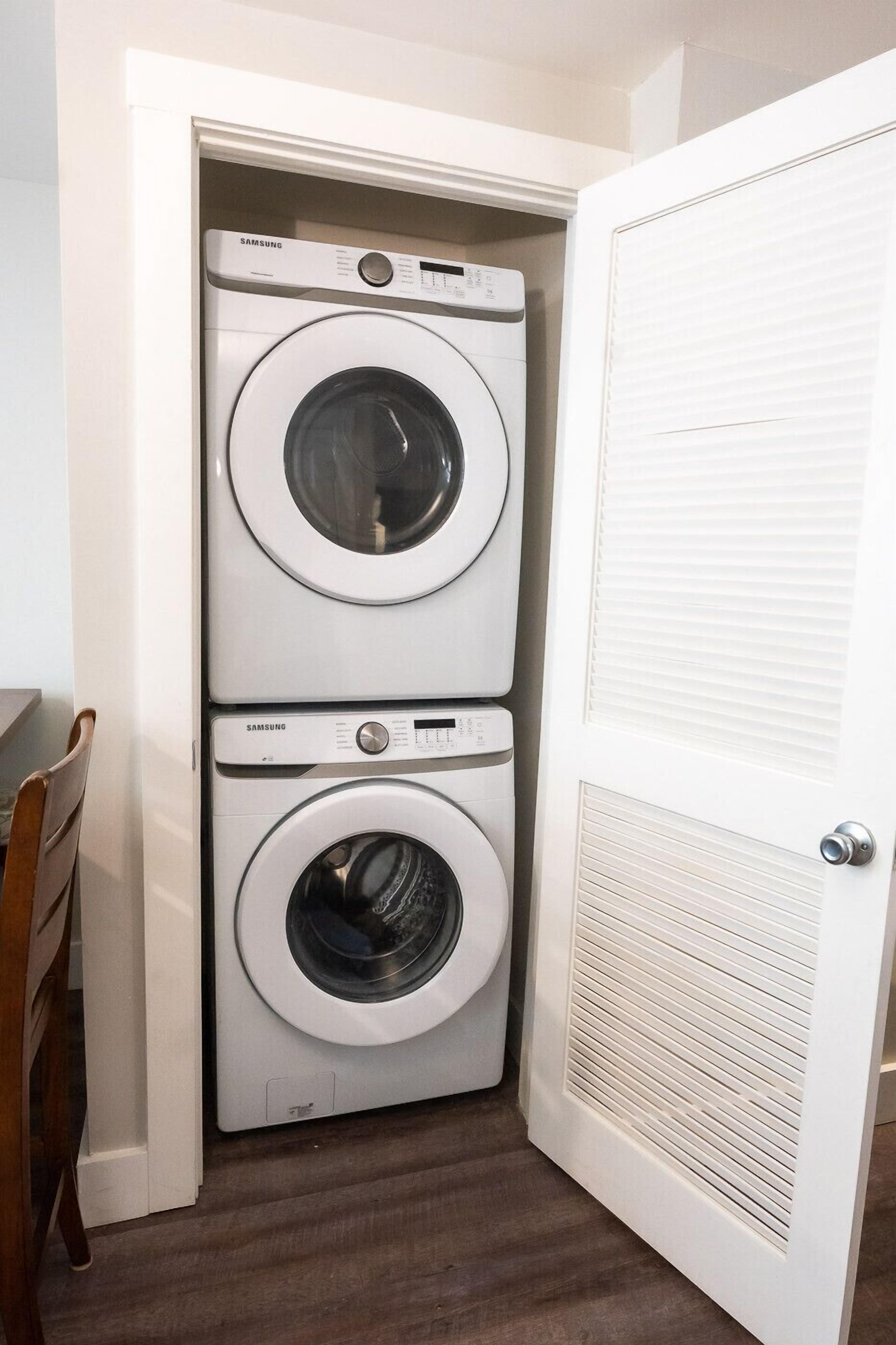A laundry room with two stacked washing machines, a wooden chair, and a white door with a vent.