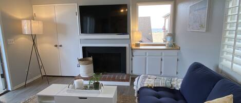 Un salon avec un canapé bleu foncé, une table basse blanche, un téléviseur à écran plat fixé au-dessus d’une cheminée et une fenêtre donnant sur la plage.