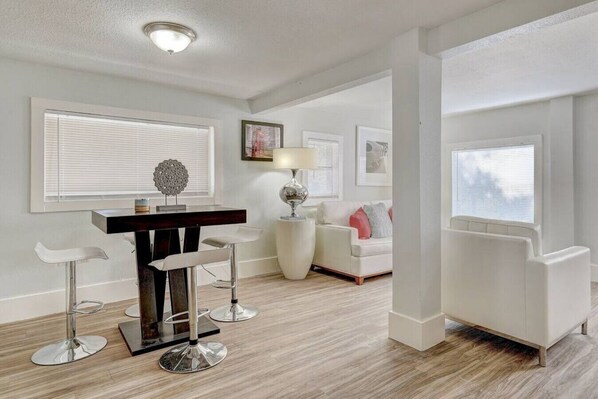 A modern living room with a dining area, featuring a wooden dining table, bar stools, a white sofa, and a floor lamp. - Downtown Round Rock Getaway Perfect for Couples (Round Rock)