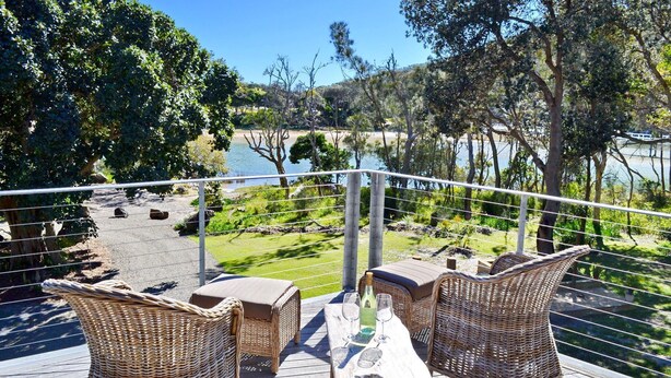 A terrace with wicker furniture, a table with glasses and a bottle, and a view of a lake and trees.