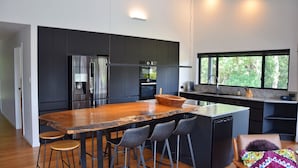 A modern kitchen with dark cabinets, stainless steel appliances, a wooden island with bar stools, and a large window offering a view of greenery.