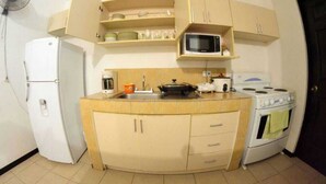 Una cocina funcional con un refrigerador blanco, microondas, estufa y lavamanos. Hay armarios sobre la encimera y un ventilador en la pared.