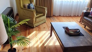 A living room with a wooden floor, a green armchair, a coffee table, a potted plant, and a large window with curtains. - Cosy Mid-Century home, Central, Off Street Parking and Fur Baby Friendly (Newington)