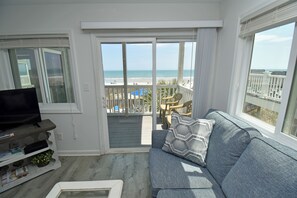 Ein Wohnzimmer mit grauem Sofa, einem Fernseher und Blick auf den Strand durch eine Glasschiebetür.