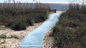 Un sentier qui mène à une plage bordée de sable et d’herbes hautes de chaque côté.