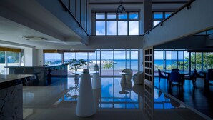 A spacious interior with large windows offering ocean views, a modern kitchen area, and a dining space with seating. - Umi No Terrace Yomitan Toya (Yomitan)