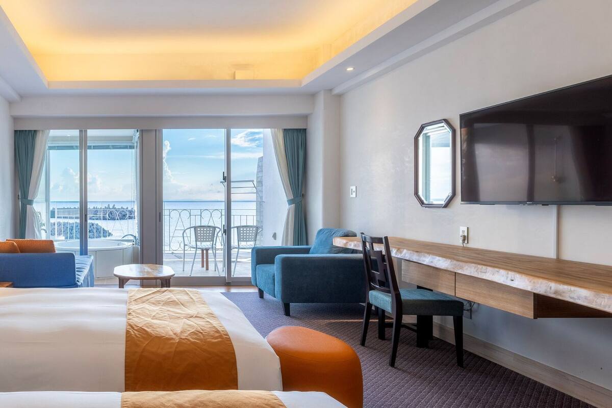 A hotel room with a large bed, a desk, a chair, and a TV. There is a balcony with a view of the city.