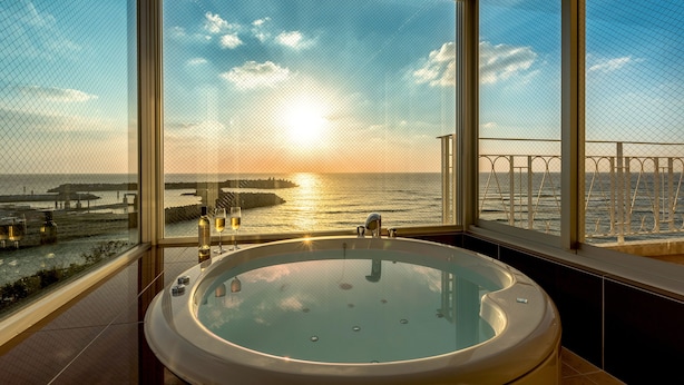 A hot tub with a view of the sunset over the ocean, a balcony with a railing, and a bottle of wine on the windowsill. - Umi No Terrace Yomitan Toya (Yomitan)