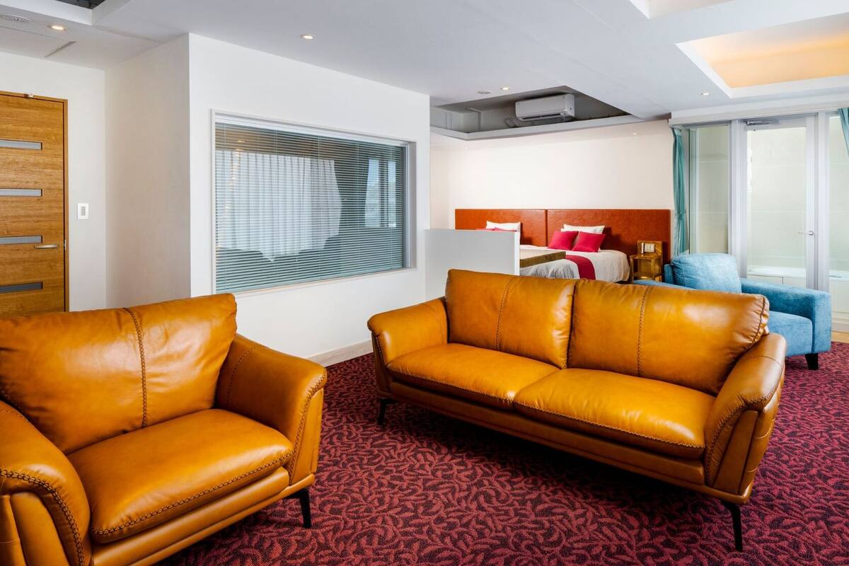 A hotel room with a brown leather sofa, a matching armchair, a bed with a red headboard, and a large window with curtains.