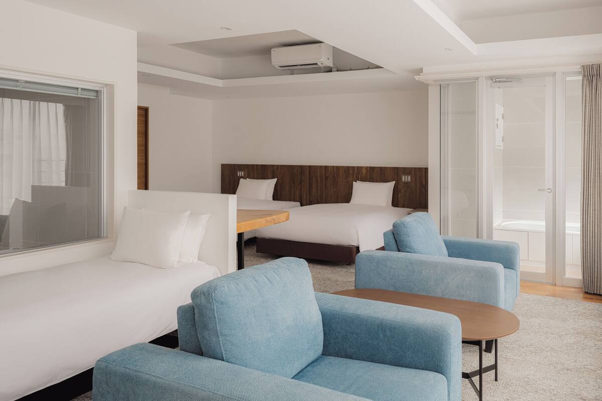 A hotel room with a bed, two blue armchairs, a small table, a window with curtains, and a sliding glass door leading to a balcony.