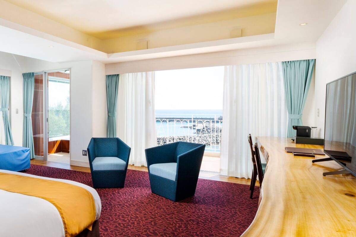 A hotel room with a bed, a blue sofa, a desk with a TV, and large windows with a view of the sea.