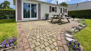 Una casa de un piso con un patio de ladrillo, una mesa de picnic y un pequeño jardín con flores moradas.