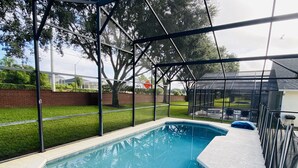 Una piscina con cerramiento de vidrio, rodeada por un muro de ladrillo y una zona ajardinada.