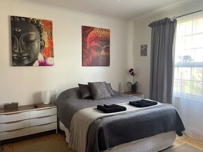 Een slaapkamer met een bed, nachtkastjes, een raam met gordijnen en twee aan de muur hangende schilderijen.