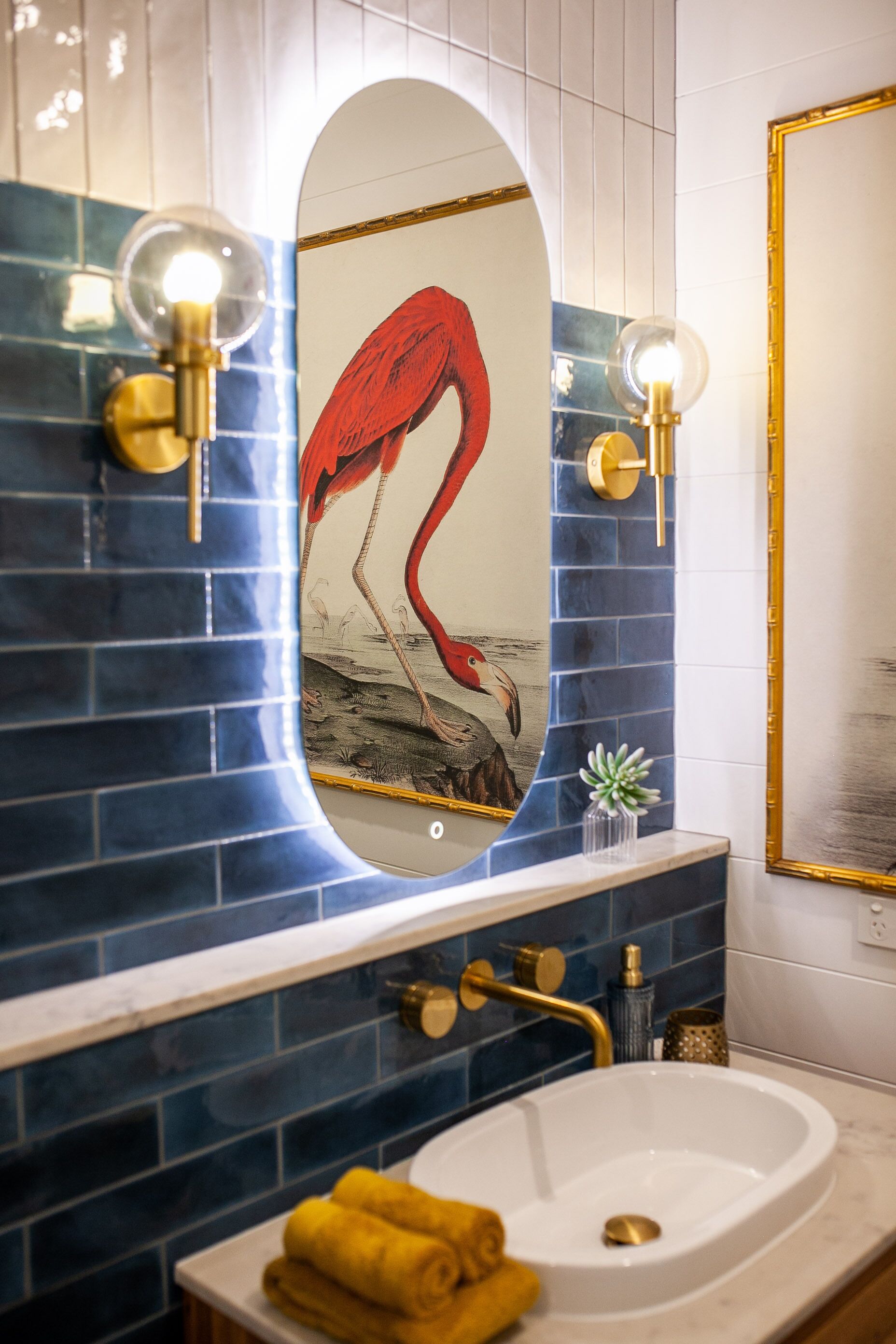 A bathroom with a white sink, blue tiled walls, and a mirror with a red bird design.