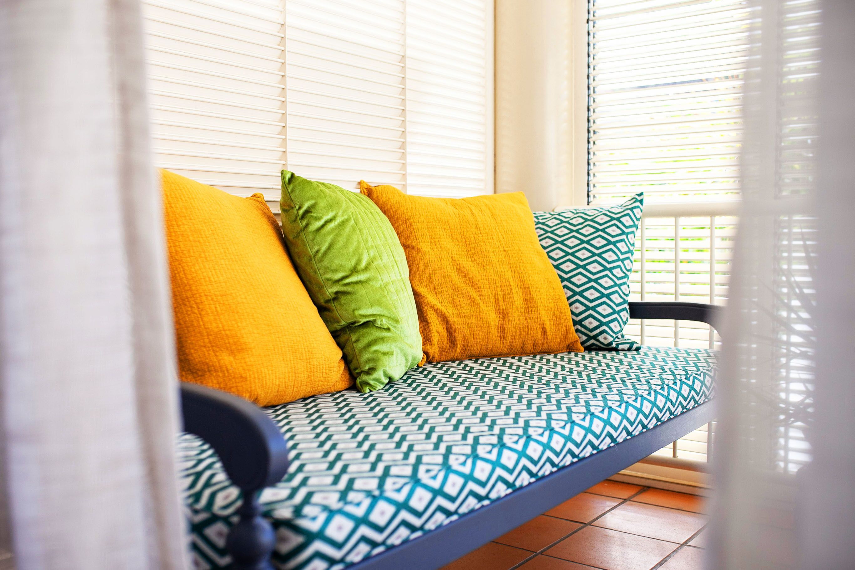 A sofa with patterned cushions and a blue frame, positioned in a room with white blinds.