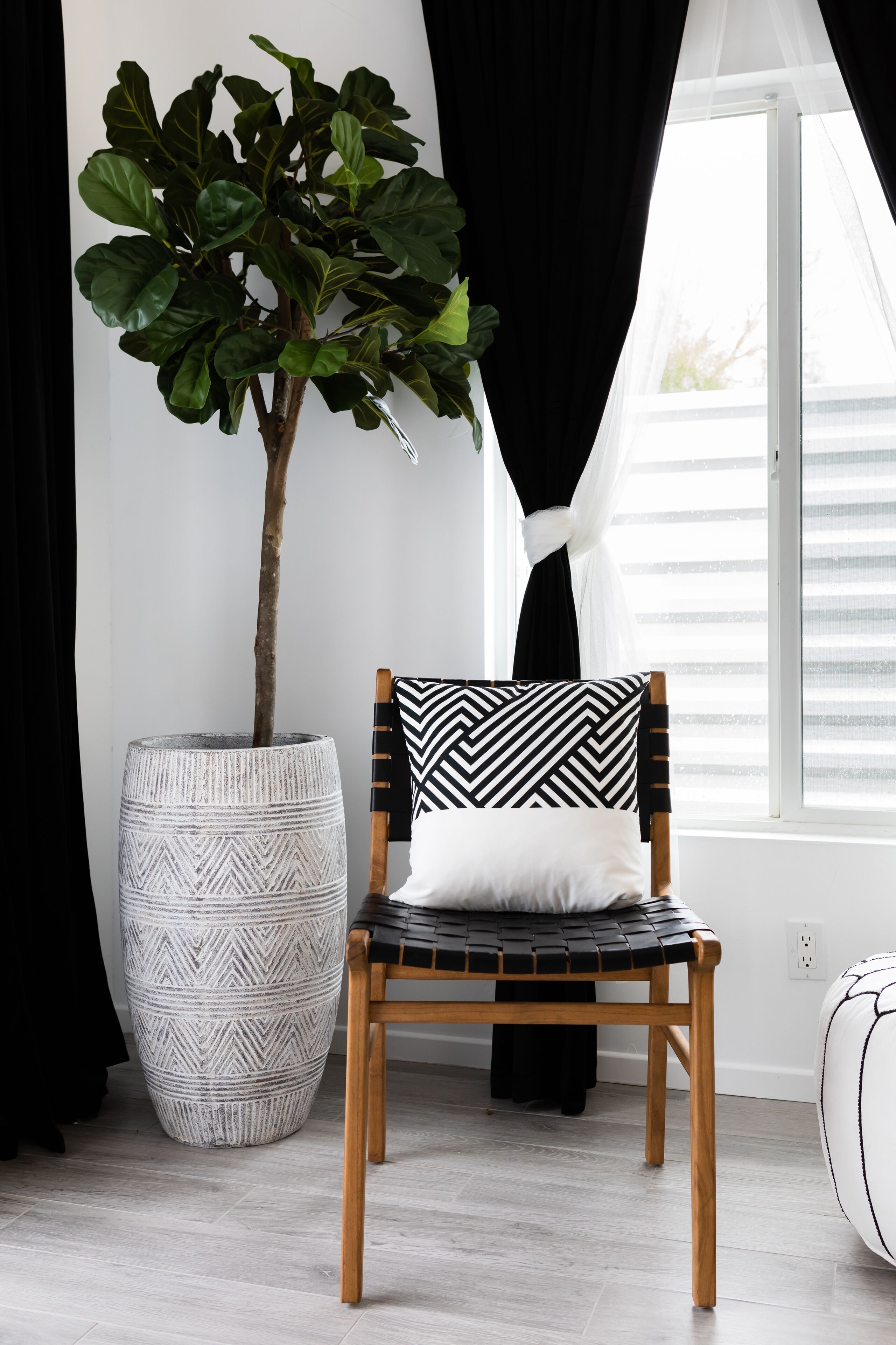 Una habitación con una planta grande en maceta, una silla de madera con un cojín estampado en blanco y negro, y una ventana con persianas.