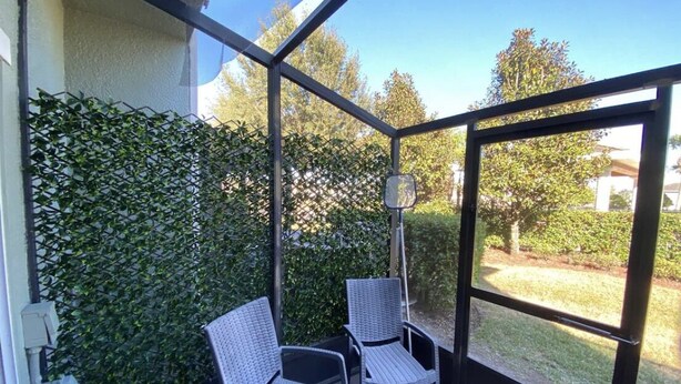 A covered patio with two wicker chairs and a view of a garden through a glass enclosure.