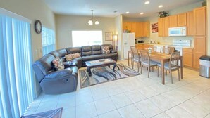 A modern living room with a sectional sofa, dining table, and chairs. There is a kitchen area with wooden cabinets and a microwave.