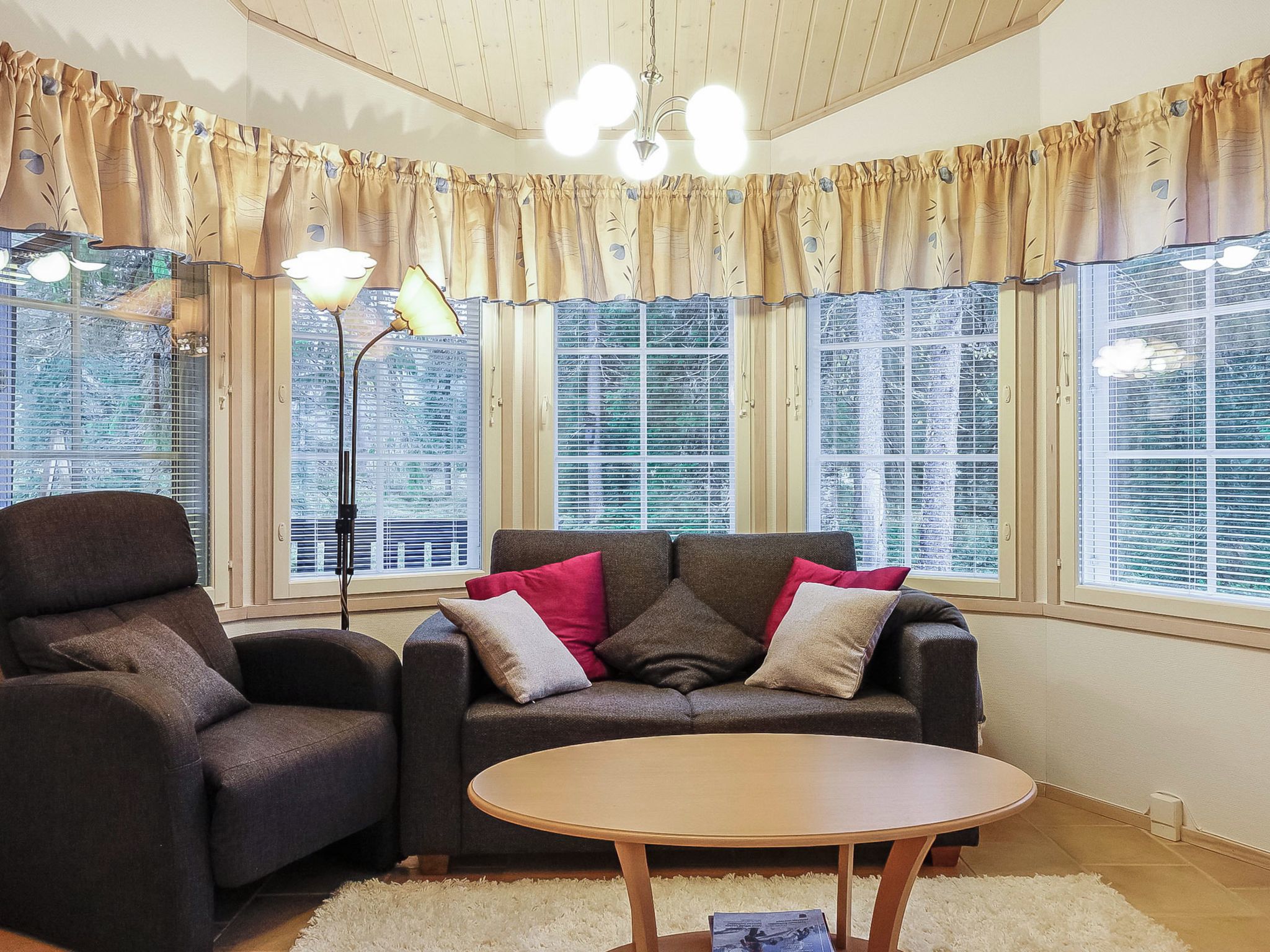 Ein Wohnzimmer mit einer Couch, einem Sessel und einem runden Couchtisch. Große Fenster mit Jalousien, eine Stehlampe und Vorhänge sind ebenfalls vorhanden.