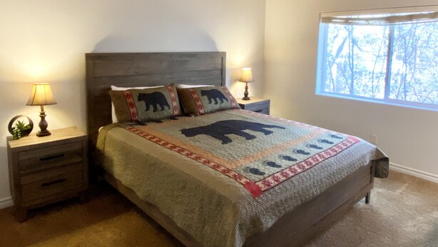 Un dormitorio con techo de madera, una cama con colcha temática de osos, dos lámparas de noche y una ventana con vistas a árboles.