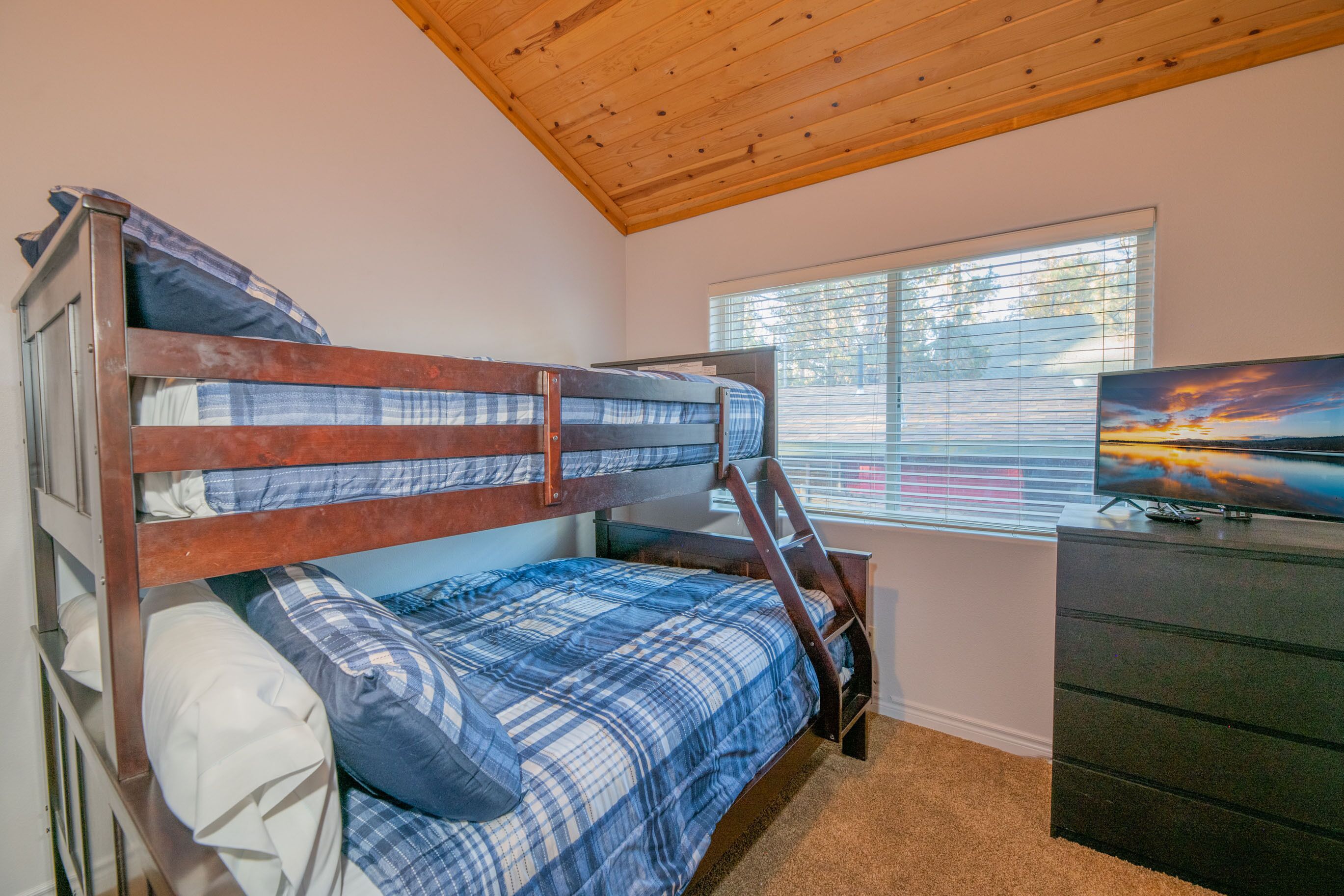 A bunk bed with a TV on a dresser in a room with a wooden ceiling and a window with blinds.