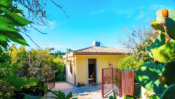 Una casa amarilla con tejado de baldosas, rodeada de follaje y cactus.