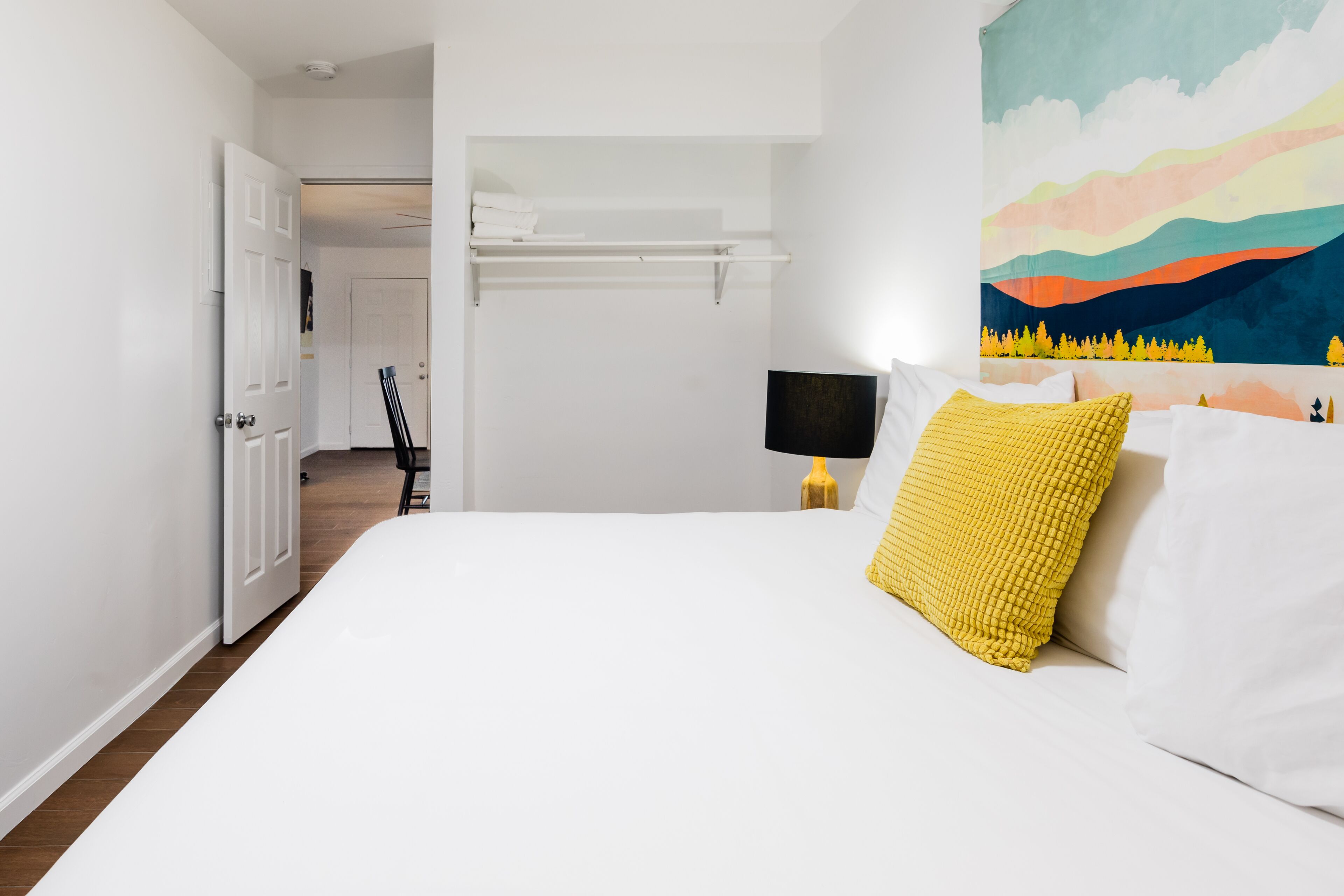 Quarto com cama grande, mesa de cabeceira, prateleira na parede e porta para outro cômodo.