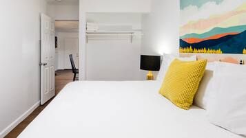 Quarto com cama grande, mesa de cabeceira, prateleira na parede e porta para outro cômodo.