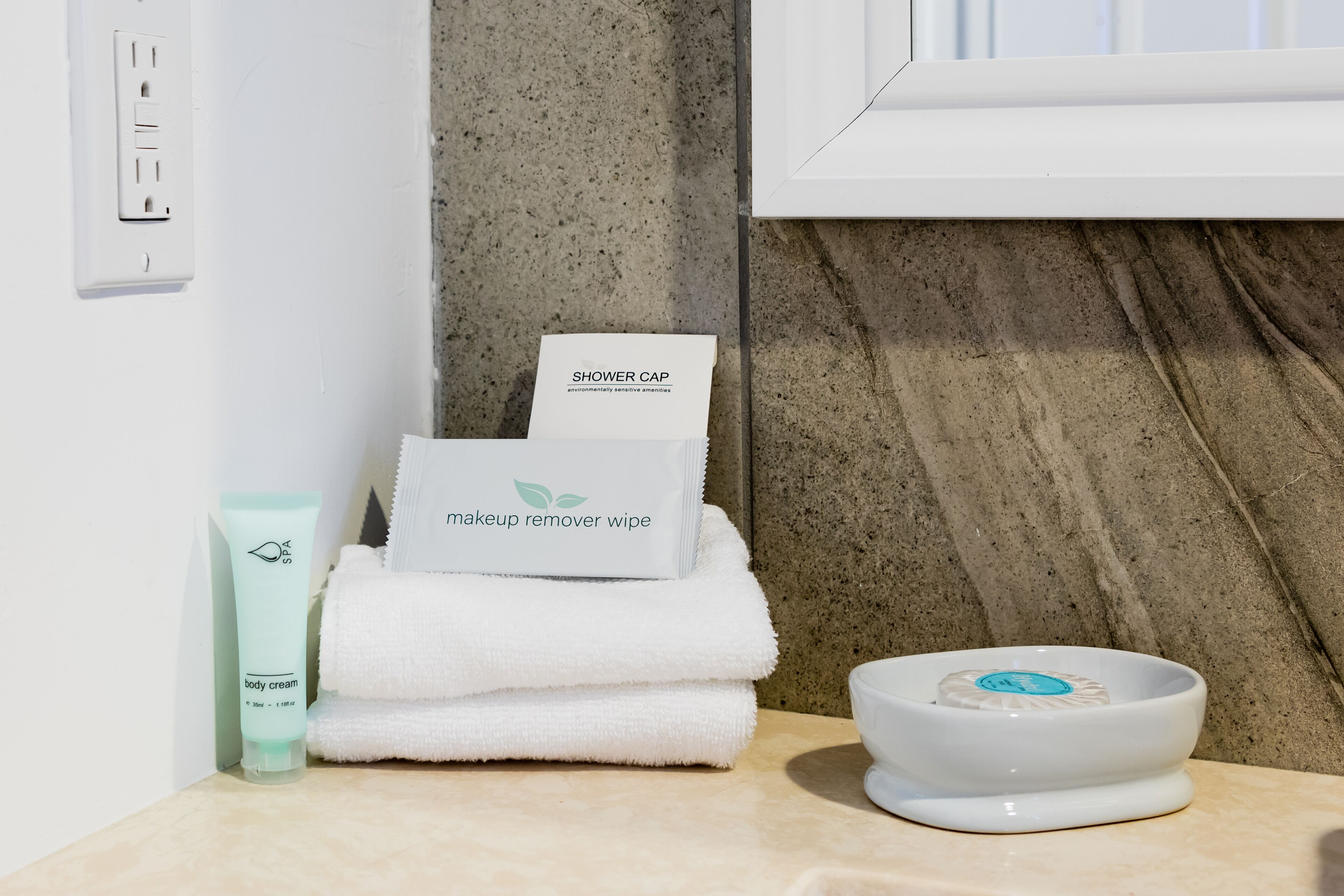 Une salle de bain avec un lavabo blanc, un porte-serviettes avec des serviettes pliées, un bonnet de douche, un lingette démaquillante et un tube de crème pour le corps.