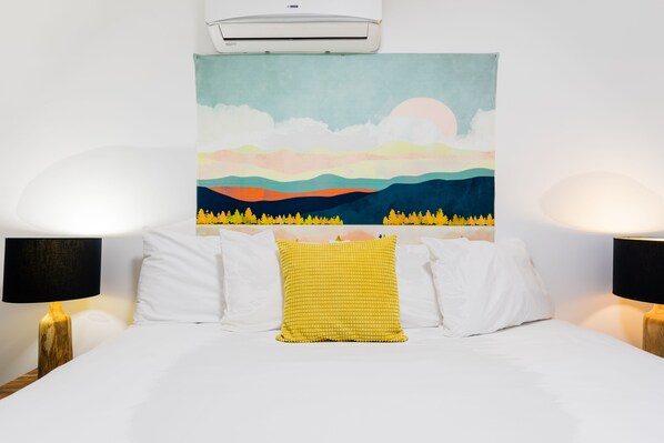 A bed with white bedding, a yellow textured pillow, and a colorful landscape painting above it.