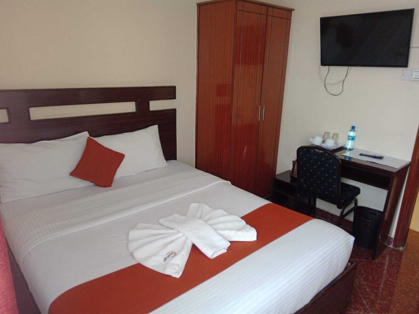 A hotel room with a bed, a wooden wardrobe, a desk with a chair, a television, and a water bottle.