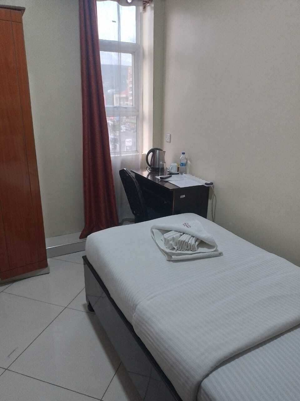 A hotel room with a bed, a desk with a kettle, a chair, and a window with curtains.