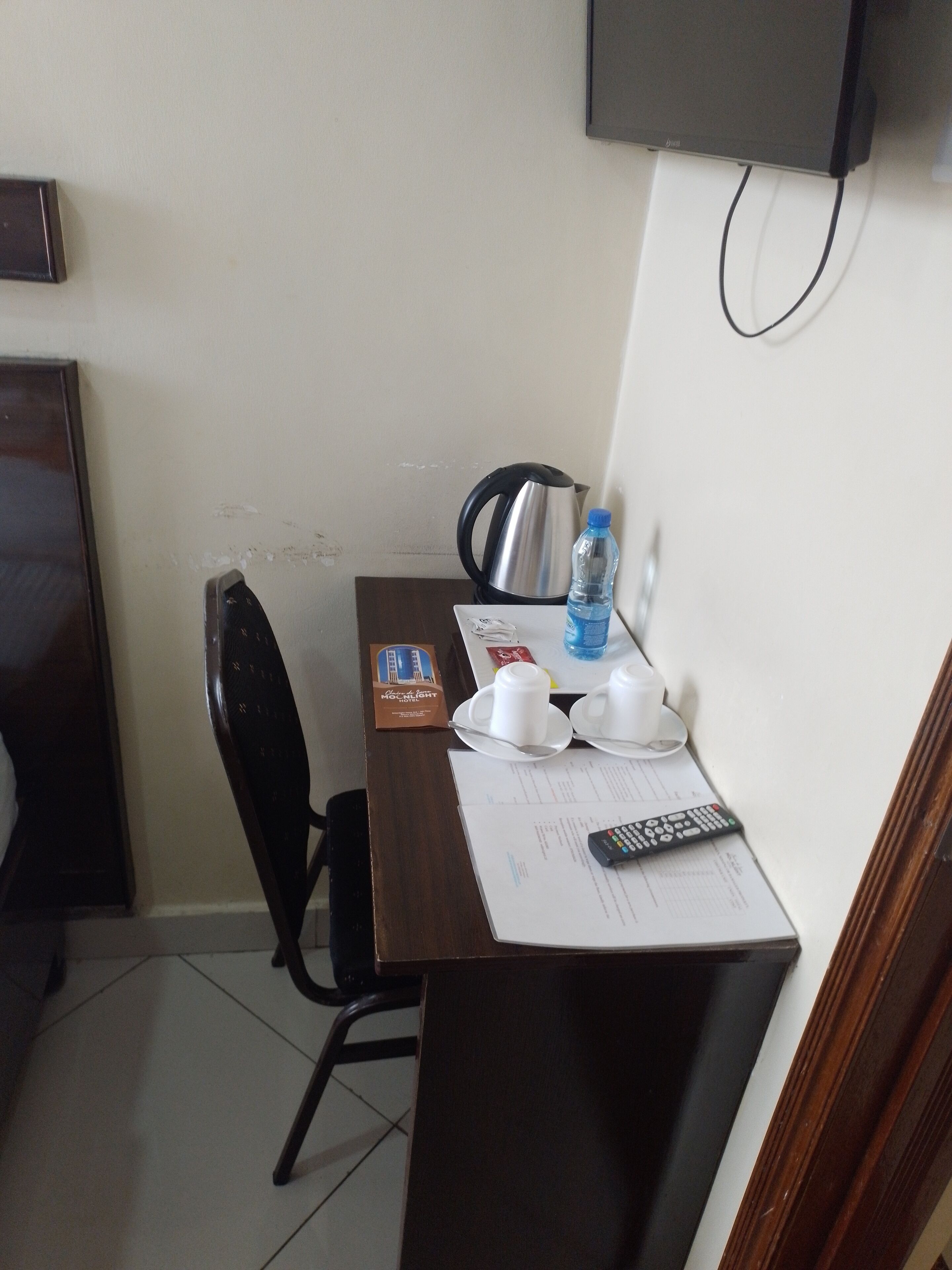 A small hotel room with a wooden desk, a kettle, a water bottle, a remote control, and a television mounted on the wall.