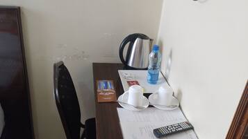 A small hotel room with a wooden desk, a kettle, a water bottle, a remote control, and a television mounted on the wall.