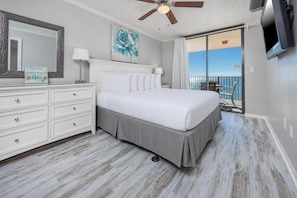 A bedroom with a bed, a dresser, a ceiling fan, a painting, and a balcony with a view.