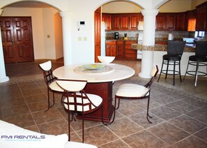 A dining area with a round table and four chairs, a kitchen in the background, and a large wooden door.