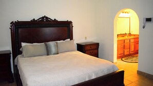 Ein Schlafzimmer mit einem hölzernen Bett, einem Nachttisch und einer Kommode. Durch einen Bogen ist ein Badezimmer zu sehen.