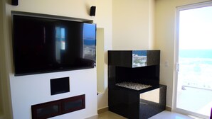Una sala de estar con un televisor de pantalla plana grande montado en la pared, un mueble de entretenimiento negro y un patio francés que da a un balcón con vista al océano.