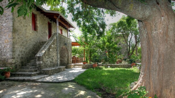 Un edificio de piedra con una escalera que lleva a una puerta de madera, rodeado por un jardín con plantas en macetas y un árbol grande.