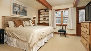 Une chambre avec un grand lit, une tête de lit en bois, une commode, une chaise, une fenêtre avec des rideaux, une étagère avec des objets décoratifs et un tableau au mur.