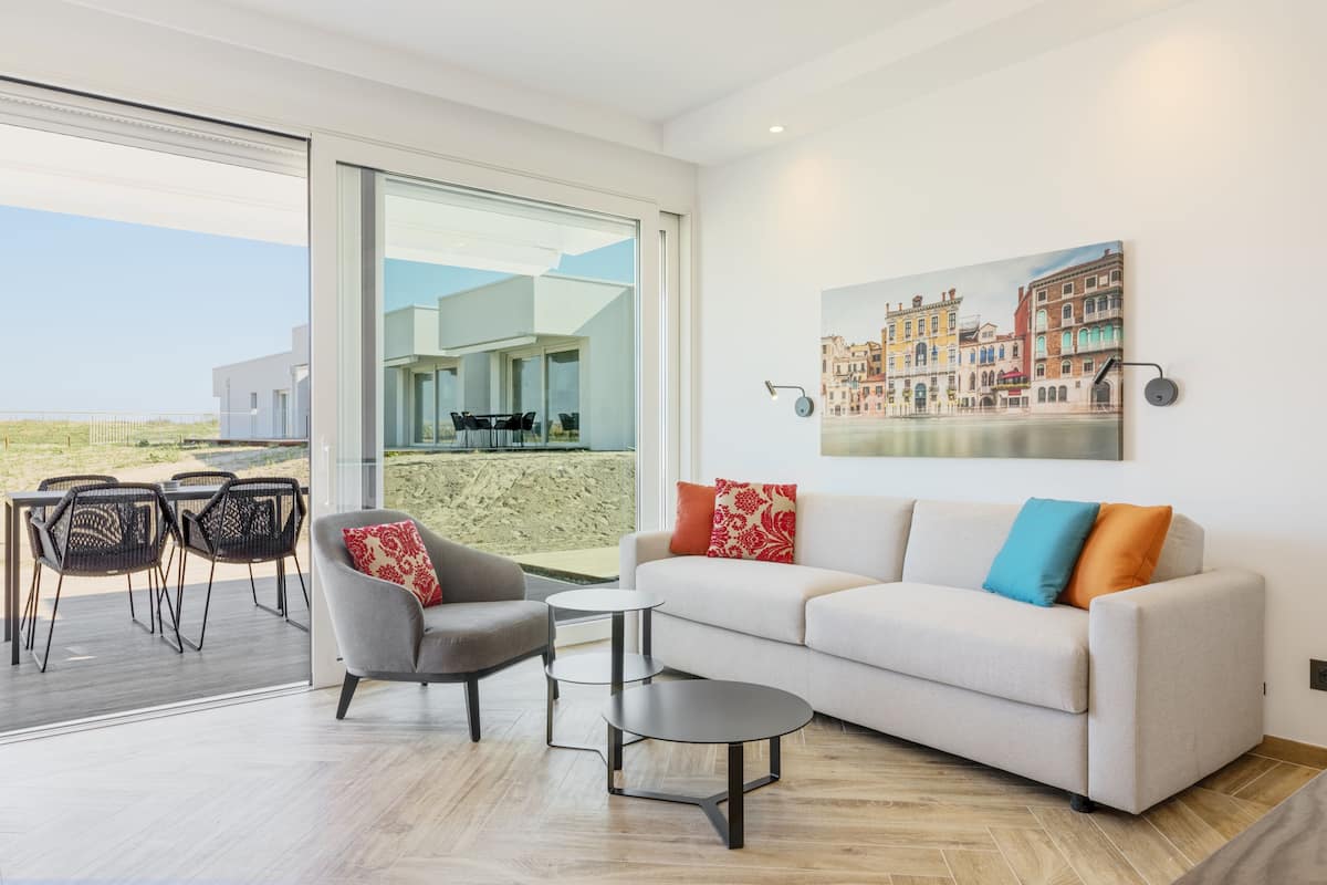 Un soggiorno moderno con divano, poltrona e tavolino, una zona pranzo con sedie e un'ampia finestra vista edifici.