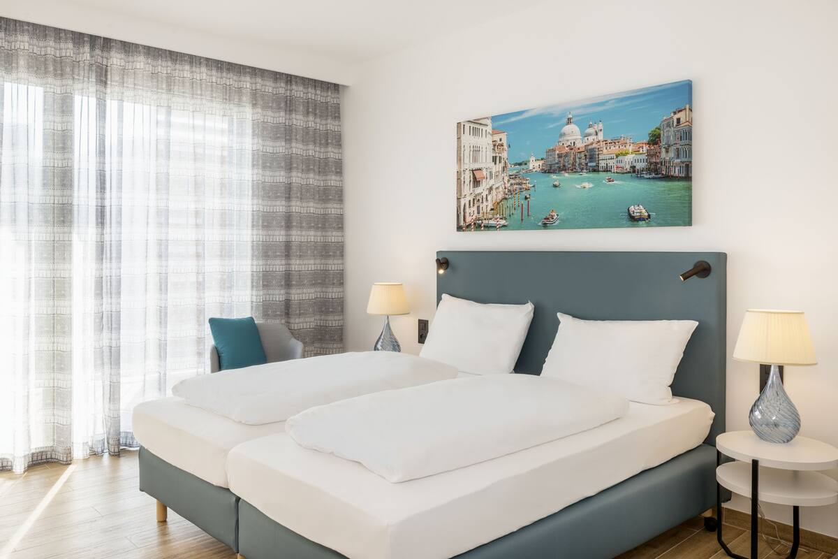 Un letto matrimoniale con lenzuola bianche, due comodini con lampade, un'ampia finestra con tende leggere e un quadro che raffigura Venezia.