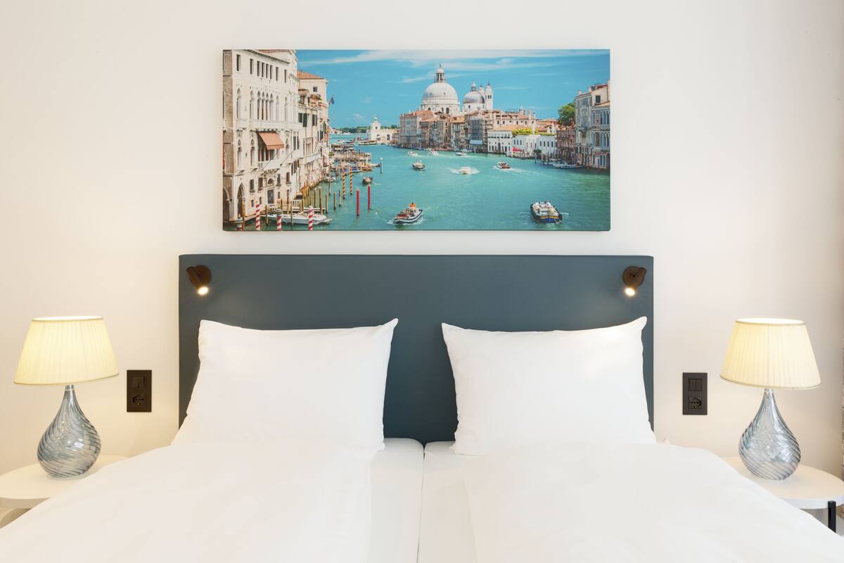 Una camera d'albergo con un letto grande, due lampade ai comodini e un quadro appeso al muro raffigurante Venezia.