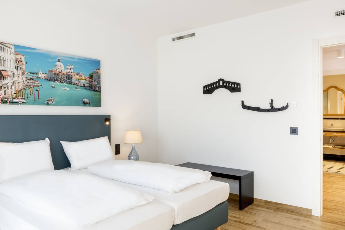 Una camera d'albergo con un letto, un comodino, una lampada e un'opera d'arte appesa al muro raffigurante Venezia.