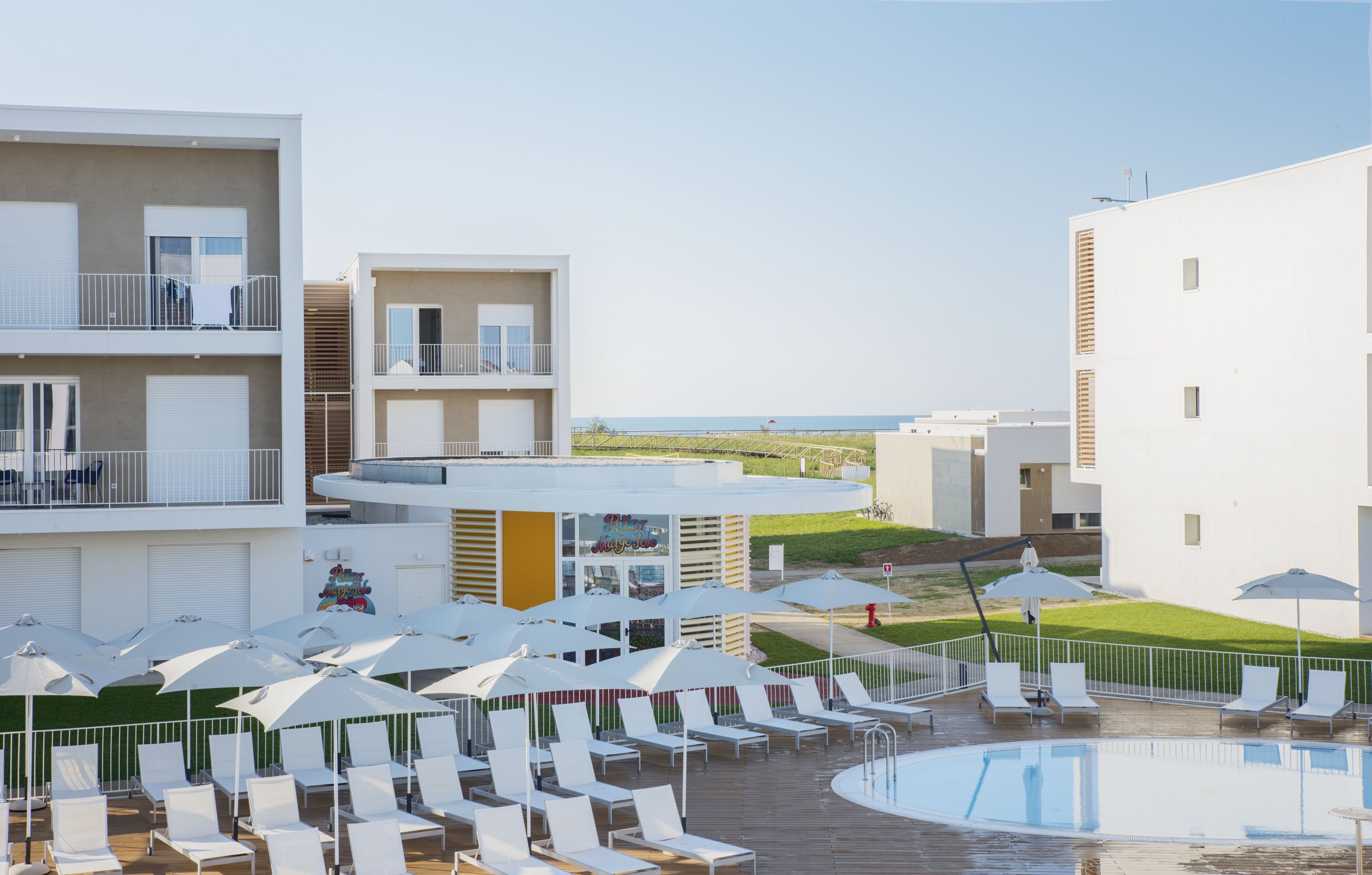 Zona junto a la piscina con sillones de descanso y sombrillas, una piscina y edificios con balcones y vistas al mar.