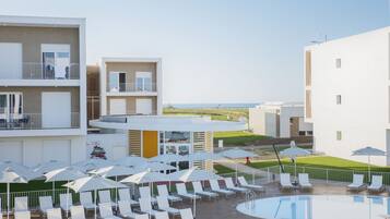 Zona junto a la piscina con sillones de descanso y sombrillas, una piscina y edificios con balcones y vistas al mar.