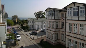 A residential area with multiple two-story houses, a street with parked cars, and a view of the sea in the background.