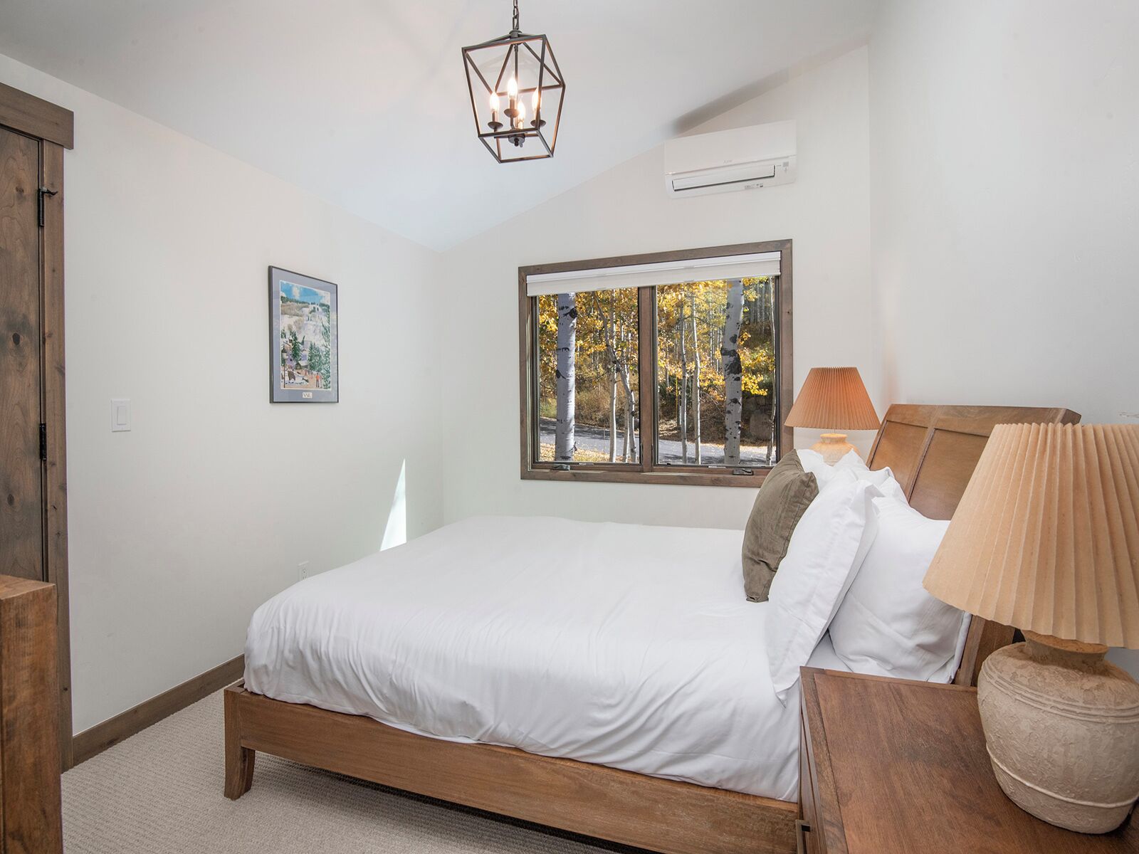 A bedroom with a wooden bed, a window with a view of trees, a wall-mounted air conditioner, and a hanging light fixture.
